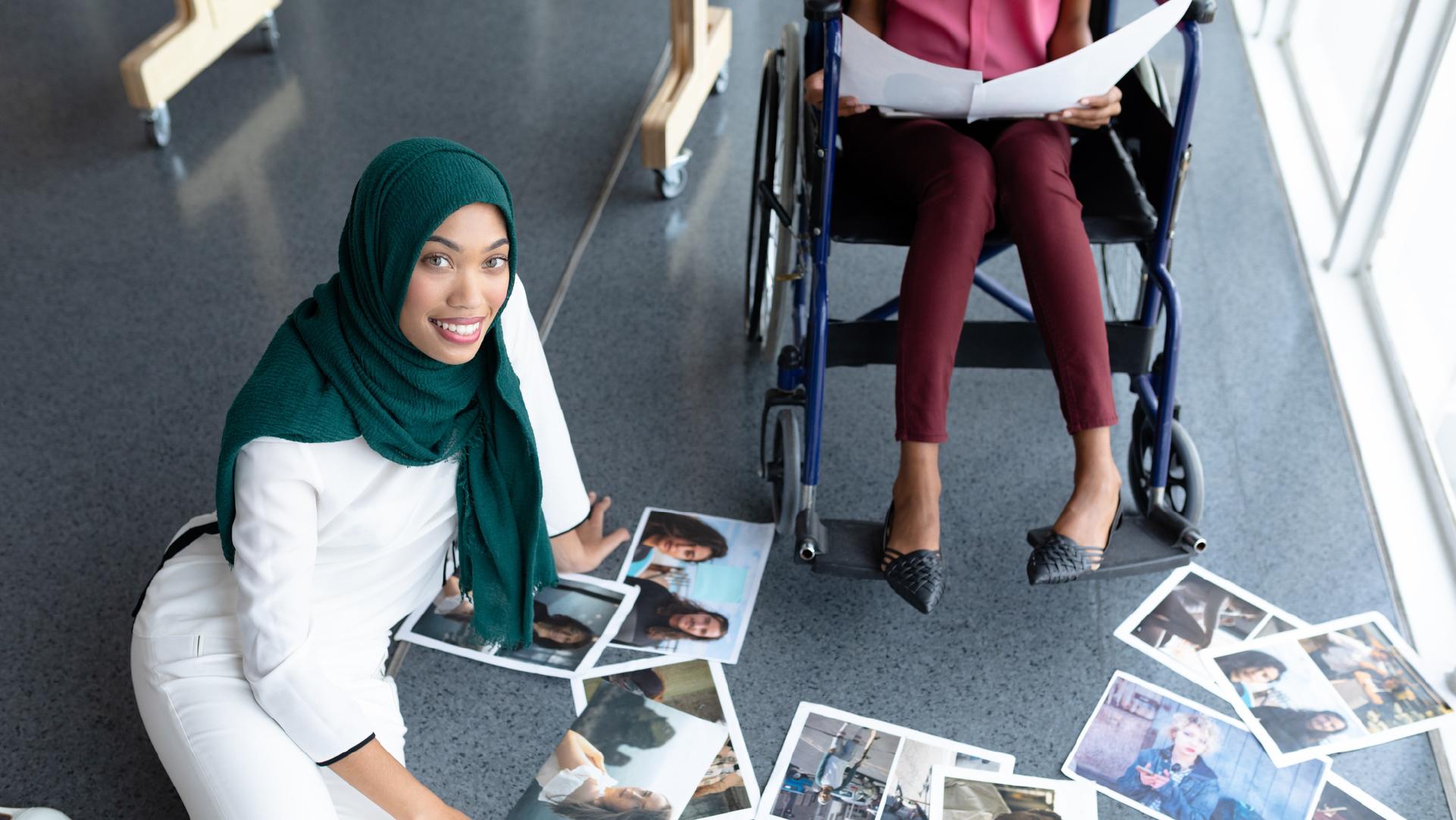 Photo of a woman sitting on the floor with some photo printouts on it, next to a wheelchair user holding two other pieces of paper