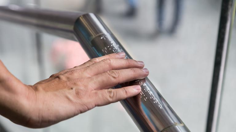 Photo of man's hand holding on to a handrail that has braille on it