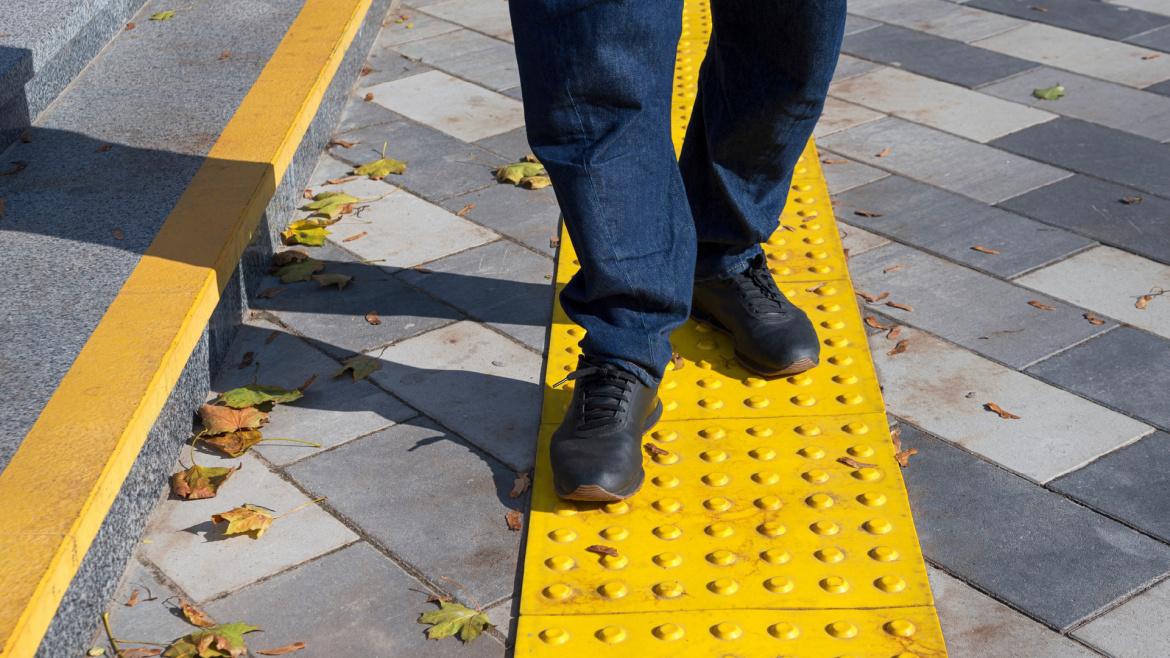Close up photo of a person's legs as they are walking on yellow tactile tiles on a pavement