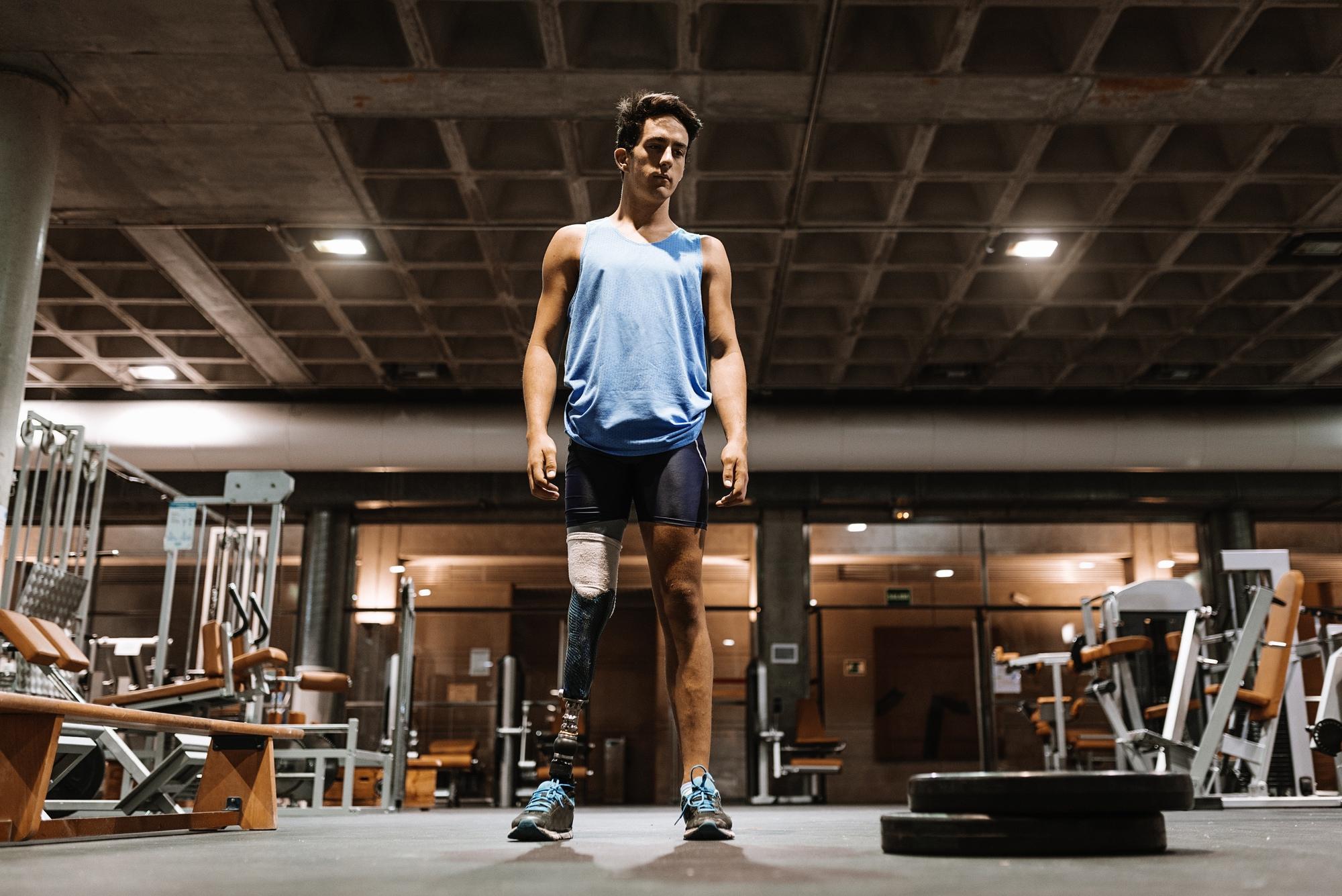 Photo of a person with a prosthetic leg, standing in a gym wearing sports shoes and surrounded by exercise equipment